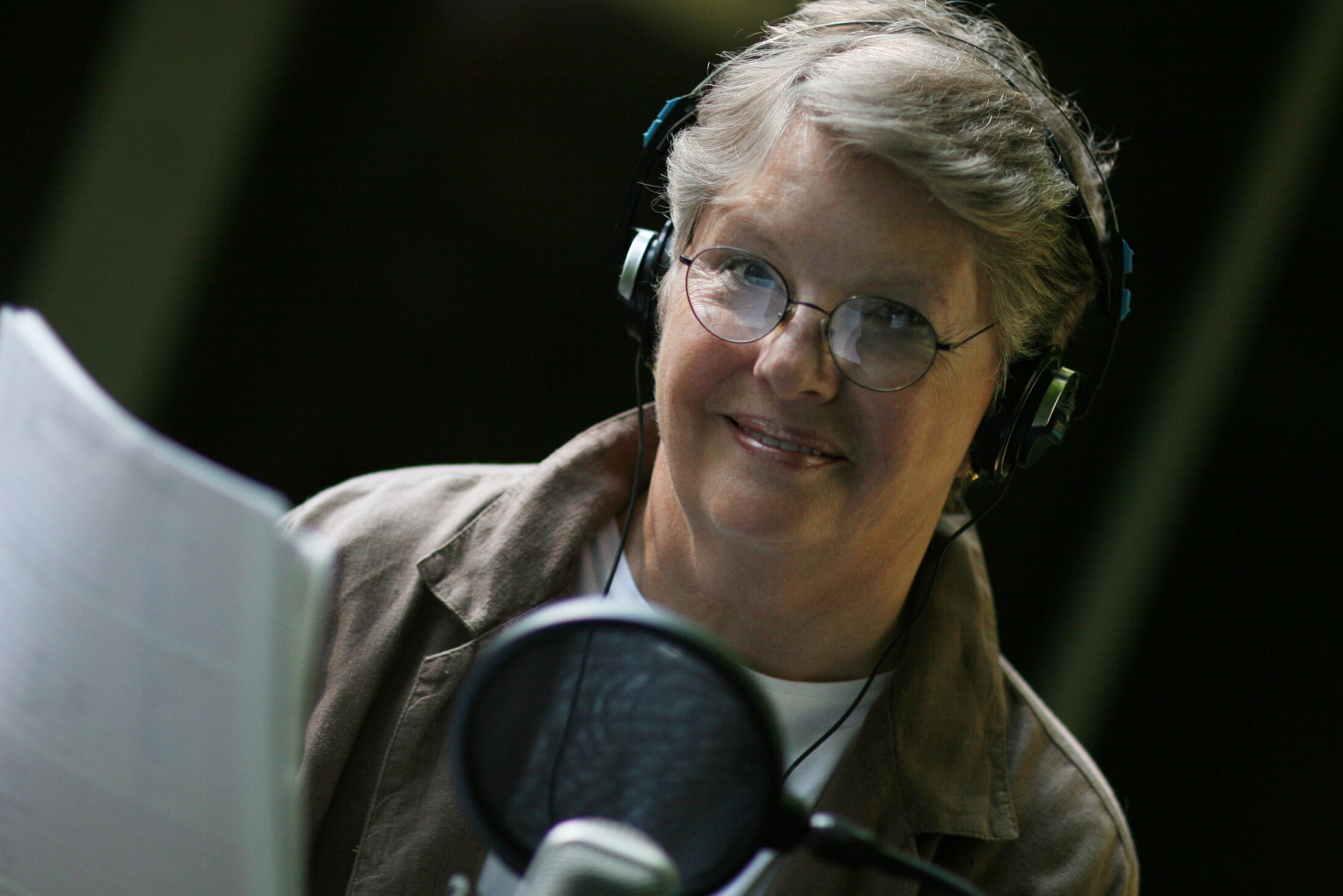 Pat Fleet in the studio, wearing headphones and reading from a script at the microphone.
